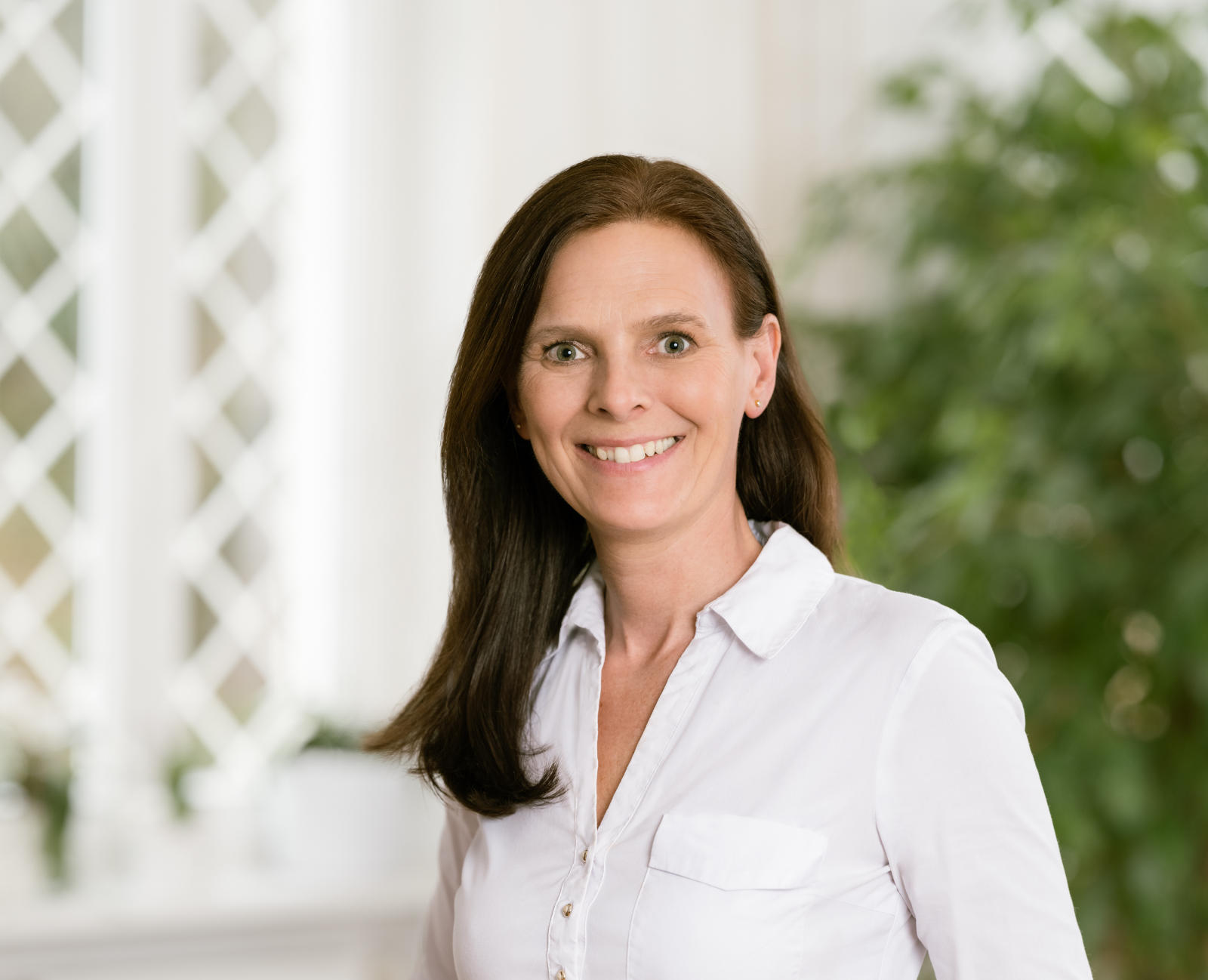 Eine lächelnde Frau mit langen, braunen Haaren und blauen Augen in einer weißen Bluse.