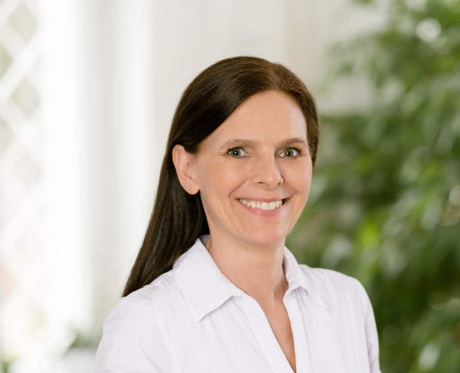 Eine lächelnde Frau mit langen, braunen Haaren und blauen Augen in einer weißen Bluse.