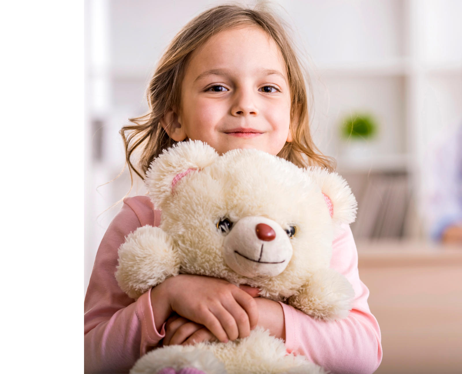 Ein kleines Mädchen drückt lächelnd einen weißen Teddy an sich. Im Hintergrund sieht man unscharf eine Ärztin.